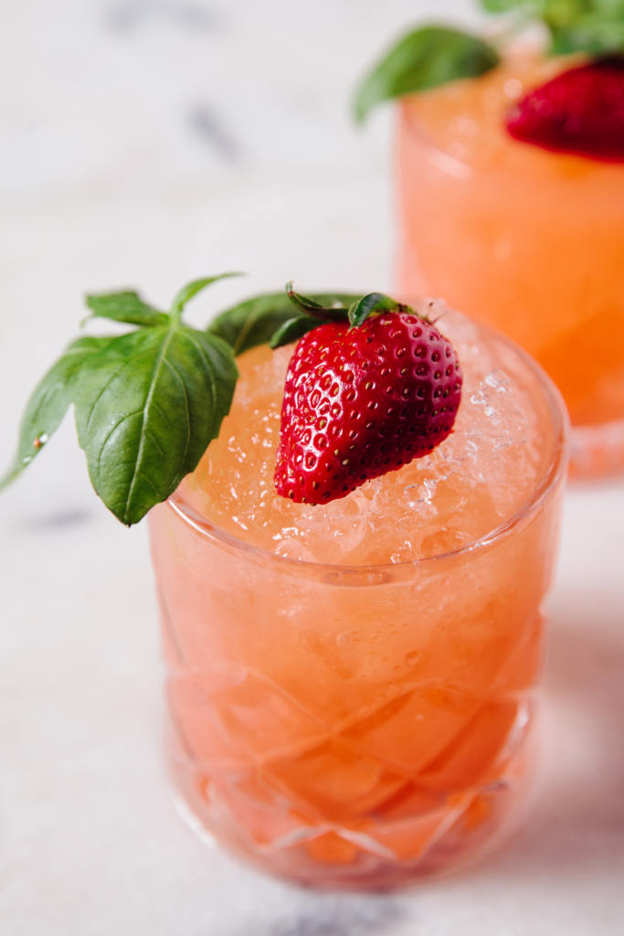 A refreshing Strawberry Basil Smash cocktail served in a textured glass filled with crushed ice. The drink has a vibrant orange-pink hue, garnished with a fresh strawberry and basil leaves.