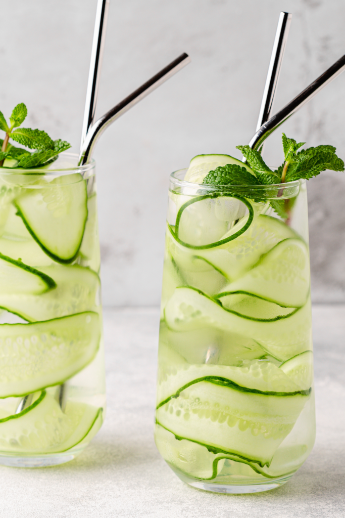 A refreshing Cucumber Mint Cooler served in a tall glass filled with ice and elegantly curled cucumber ribbons. The drink is garnished with a sprig of fresh mint, adding a vibrant green contrast.