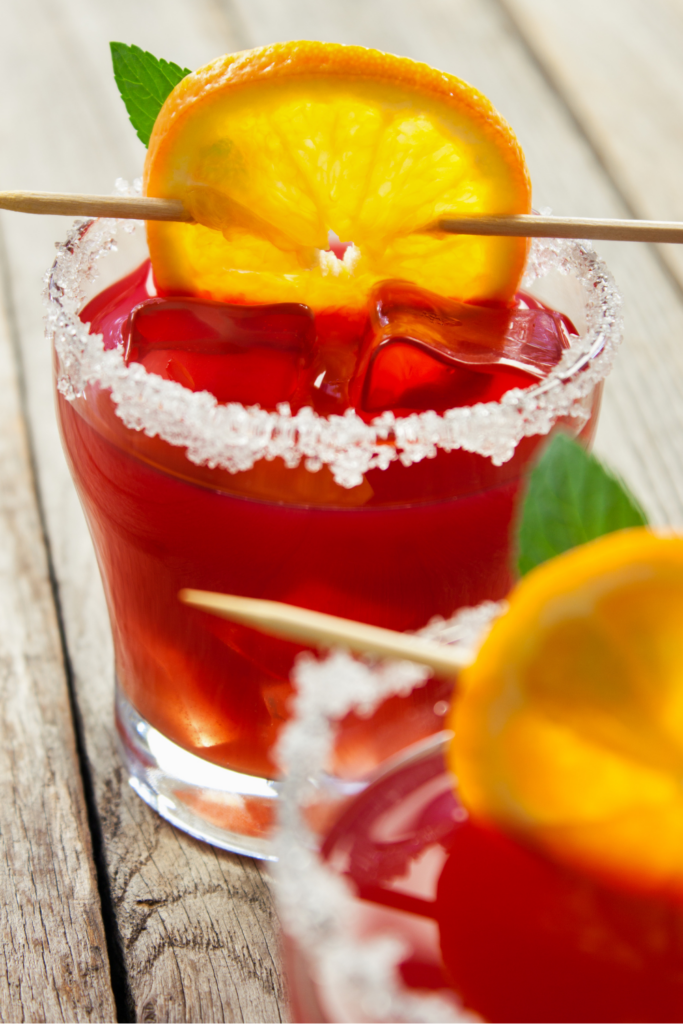 A bold and vibrant Blood Orange Margarita served in a short glass with a salted rim and large ice cubes. The drink has a deep ruby-red hue, highlighting the rich citrus flavors. It is garnished with a caramelized orange slice skewered on a wooden pick and a sprig of fresh mint